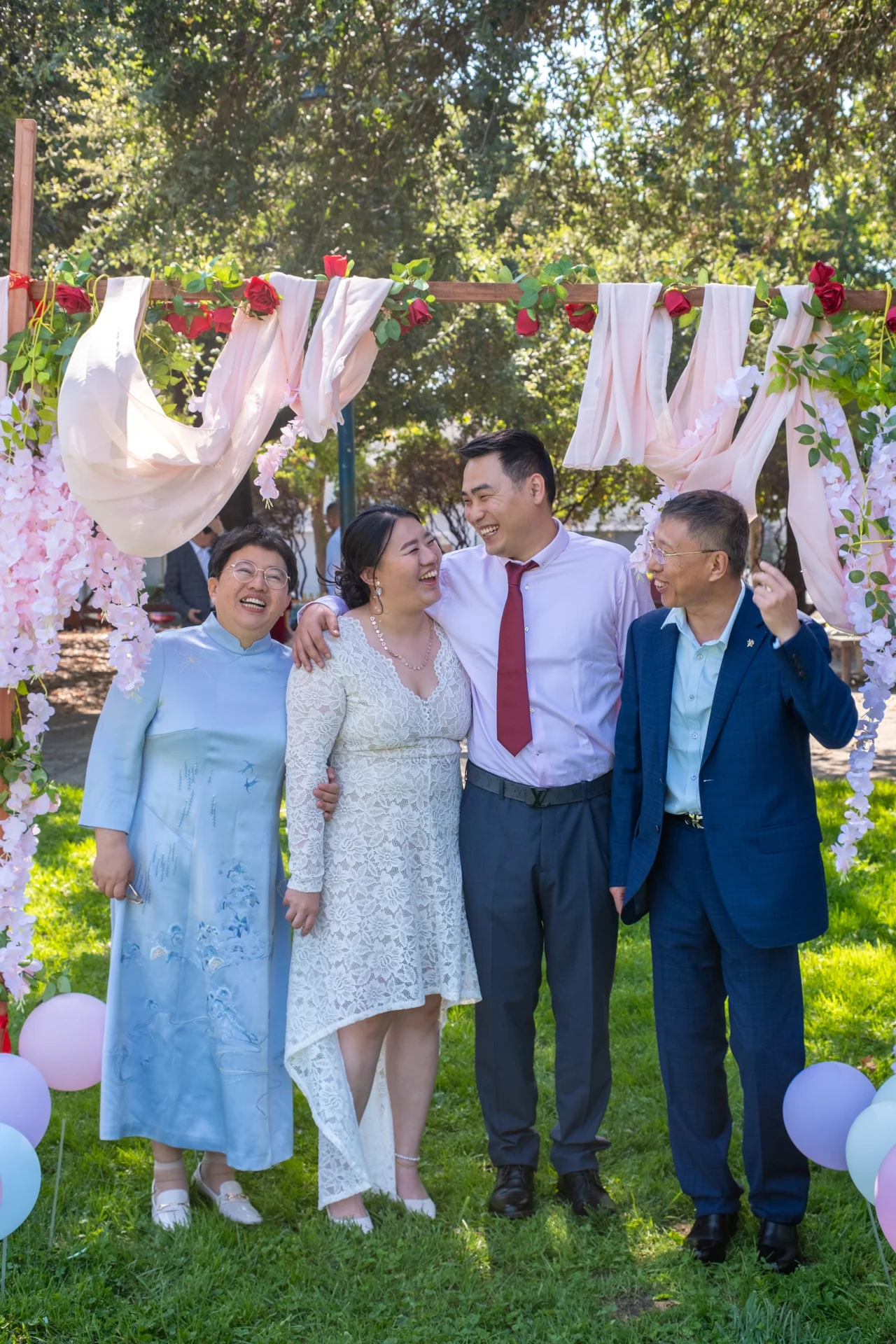 Four people laughing together beneath a flower-decorated wedding arch.