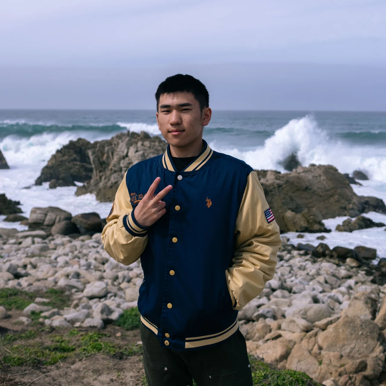 Man in a blue and gold varsity jacket beside a rocky shoreline.