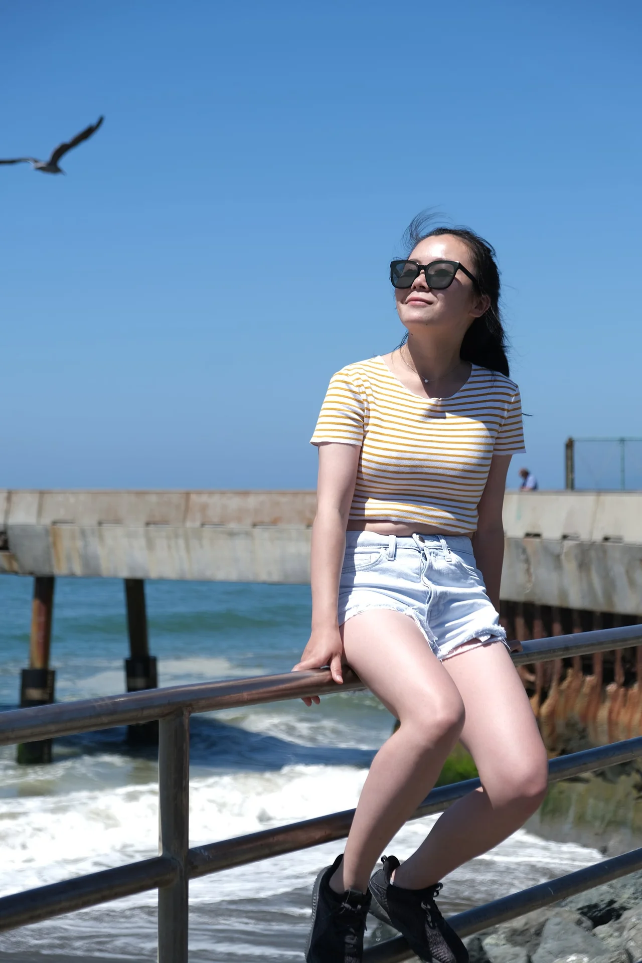 Woman in sunglasses seated on a railing beside the sea.