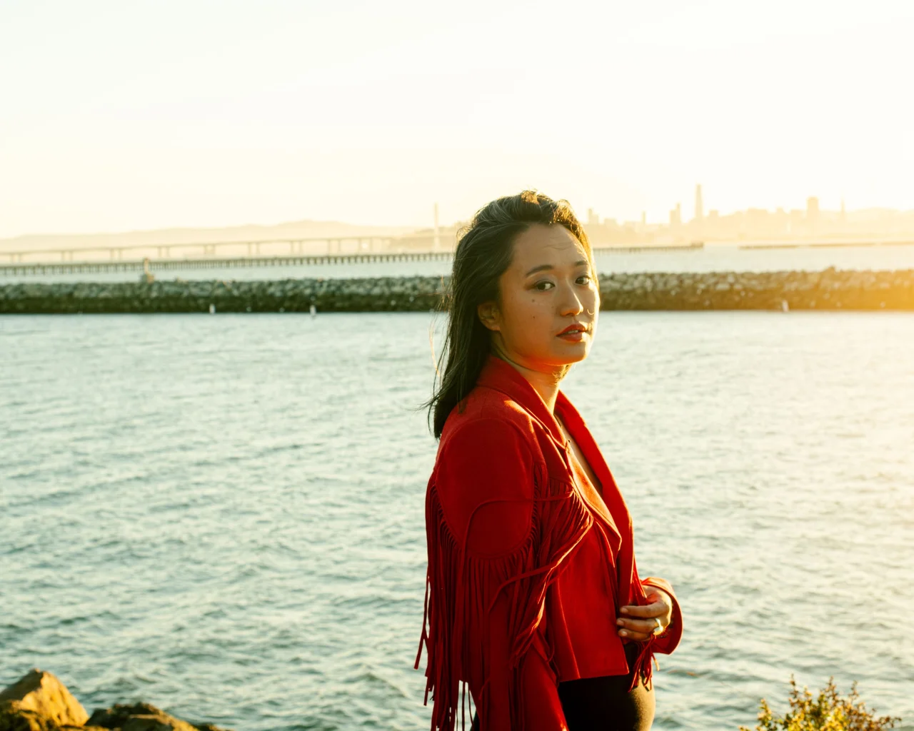 Woman in a red jacket beside the water in golden evening light.