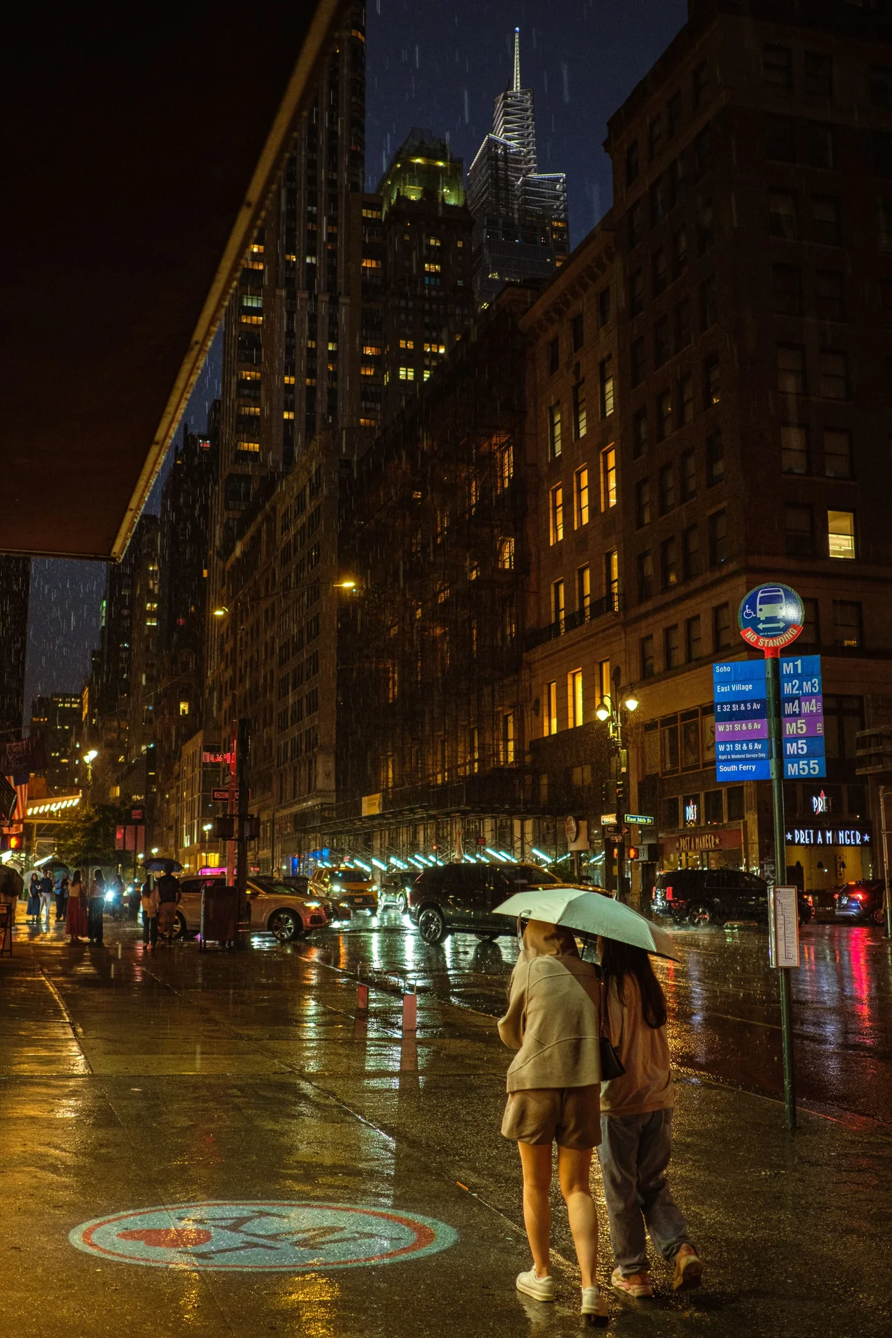 Two people sharing an umbrella on a wet city sidewalk beneath illuminated buildings.