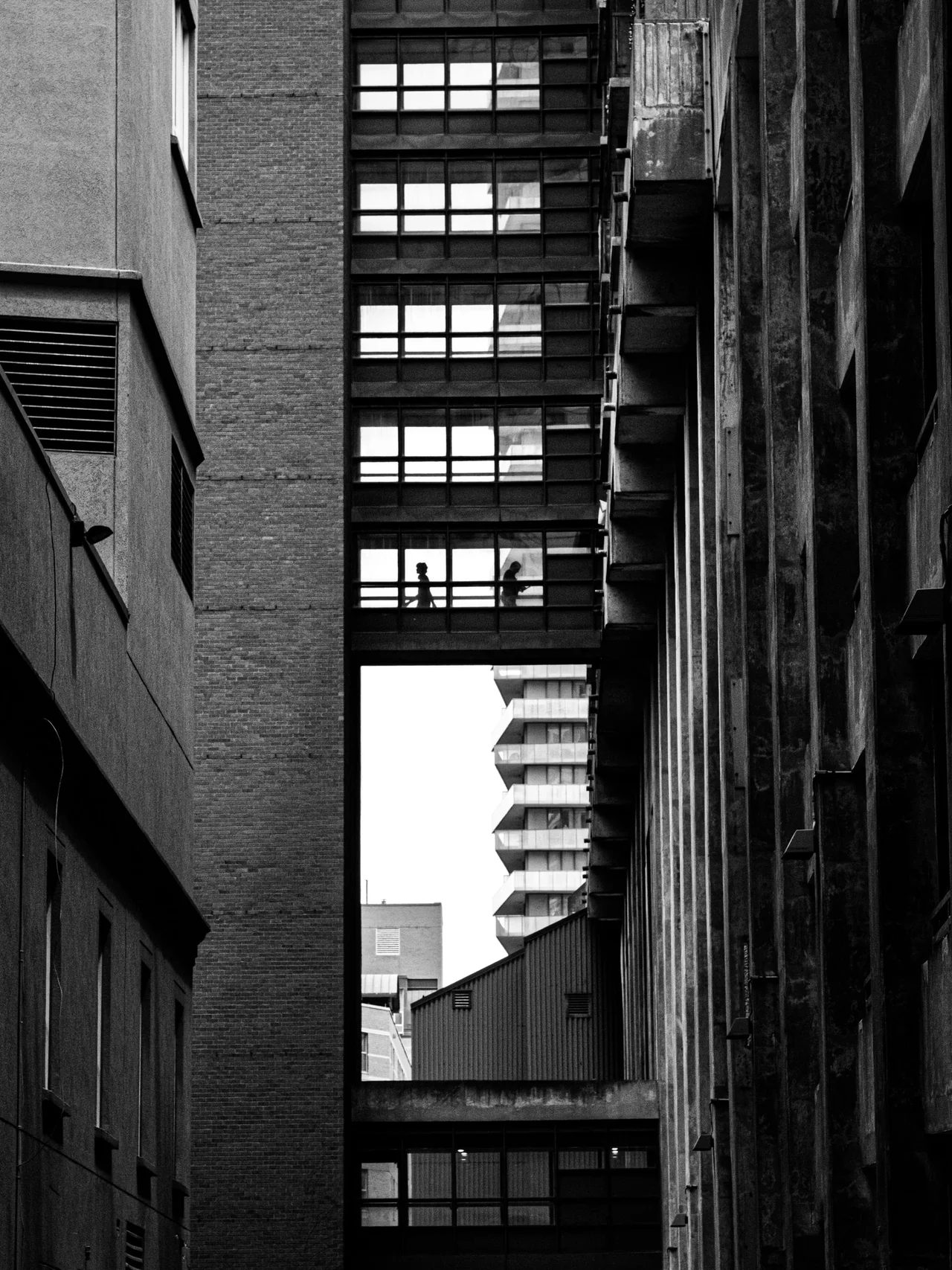 Two silhouettes inside a glass walkway between tall buildings in black and white.